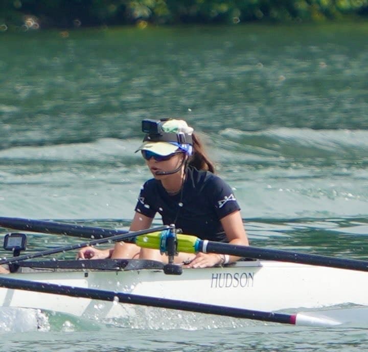 A cox in a visor and headset, seated in a white boat on the water.