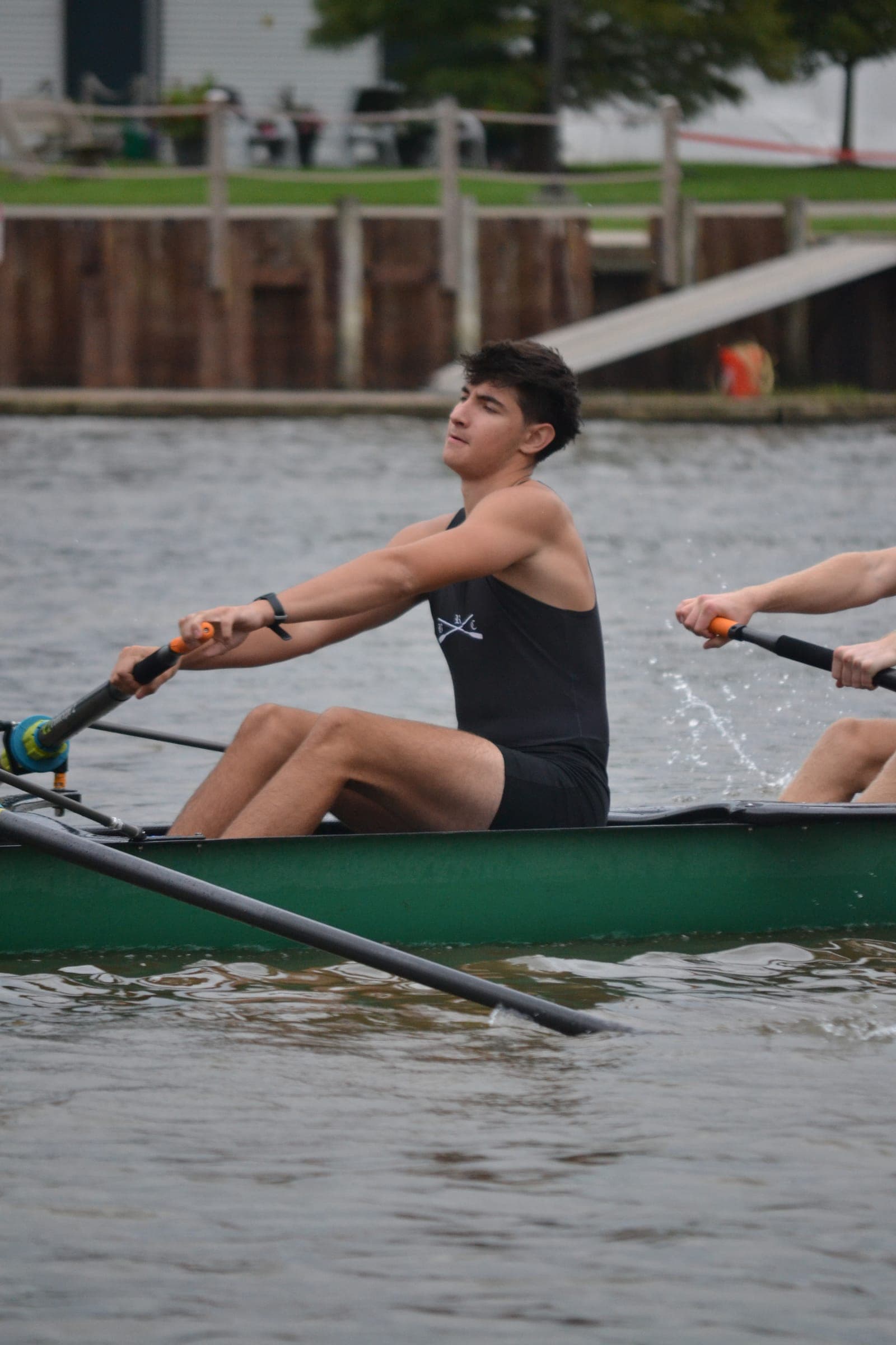 A rower in a black uniform, oar in both hands, seated in a green boat on the water.